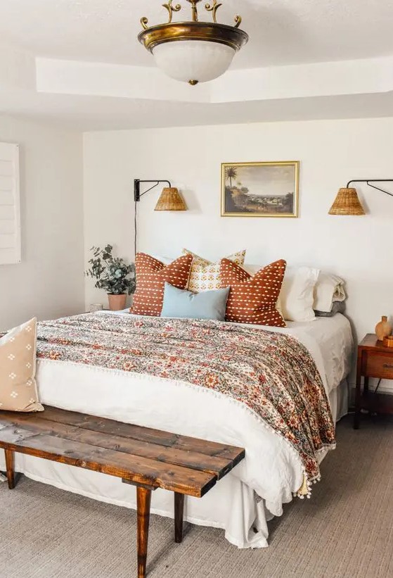 a welcoming bedroom in neutrals with a bed with floral and other printed bedding, a stained bench, a chandelier, stained nightstands