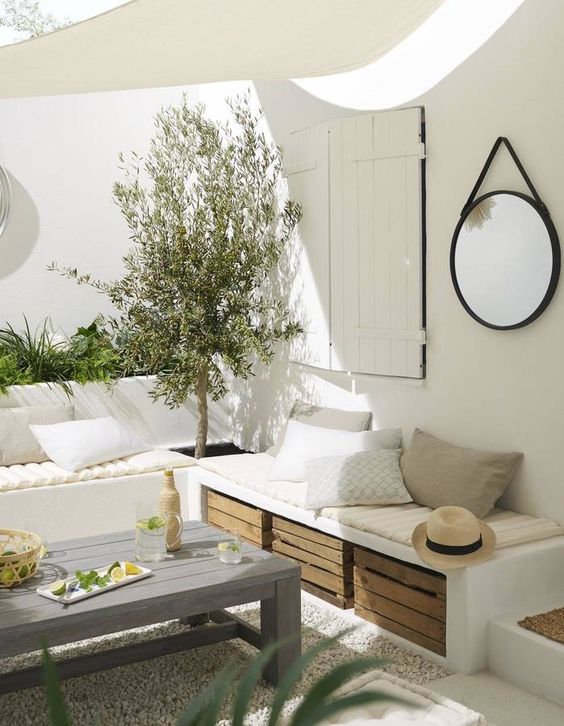 a white Mediterranean terrace with built-in benches, greenery and a tree, crates for storage and a small roof
