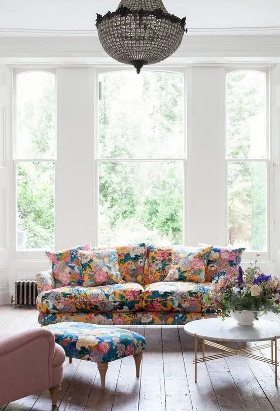 an airy living room with large windows, a floral sofa and a footrest, a pink chair, a coffee table and a crystal chandelier