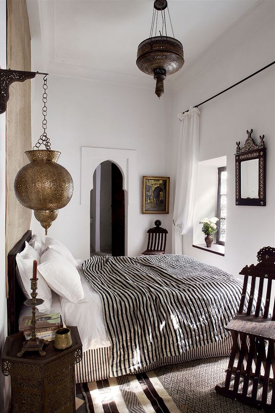 an eclectic bedroom with Moroccan influence, with a striped bed and blanket, metal Moroccan lamps and carved wooden furniture