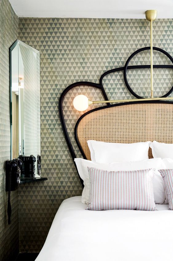 an eclectic bedroom with geometric wallpaper walls, a corner mirror with a shelf, a bed with a cane headboard and neutral bedding