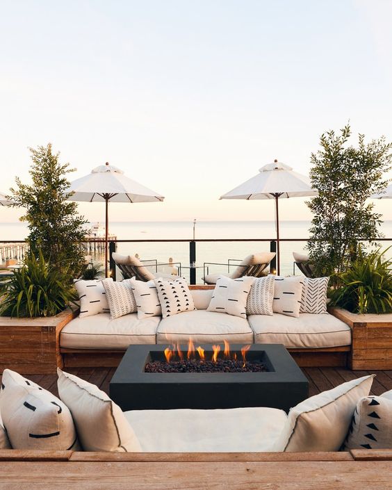 an elegant and welcoming summer terrace with a sea view, built-in sofas, a fireplace, potted greenery and trees