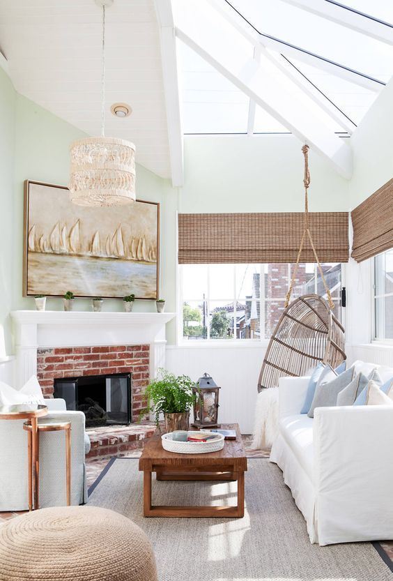 an elegant coastal sunroom with white and light blue furniture, woven shades, a brick fireplace, a woven lamp and a coastal artwork