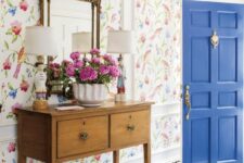 an elegant farmhouse entryway with bright floral wallpaper, a stained console table, a grene pouf, a crystal chandelier and a refined mirror