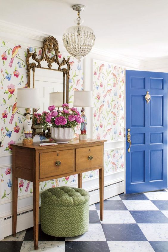 an elegant farmhouse entryway with bright floral wallpaper, a stained console table, a grene pouf, a crystal chandelier and a refined mirror