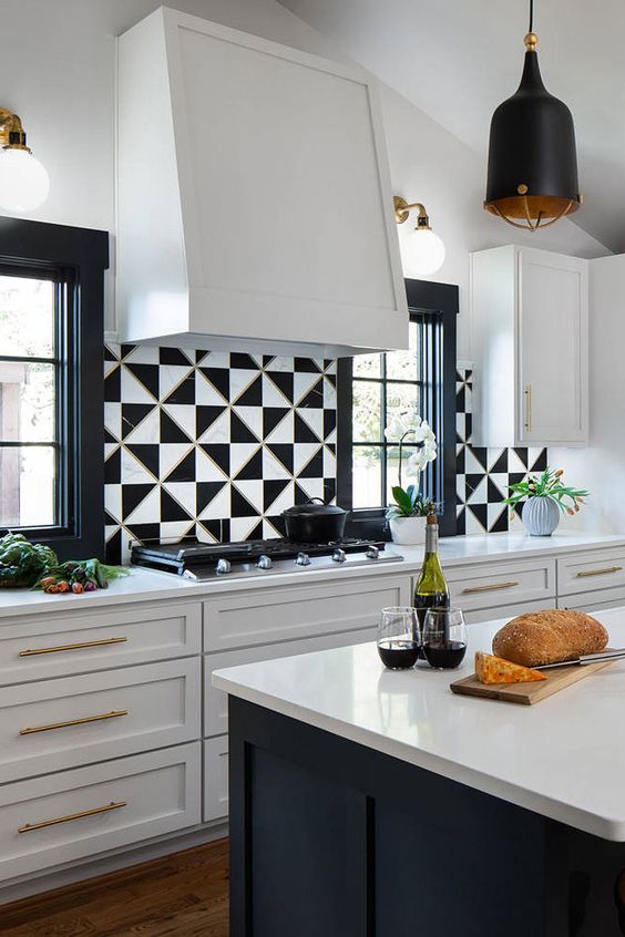 an elegant white kitchen with shaker style cabinets, a black kitchen island with white stone countertops, black and white geo tiles and a large hood