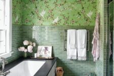an eye-catchy green bathroom with floral wallpaper and green tiles, a bathtub with paneling, a bead chandelier and some blooms