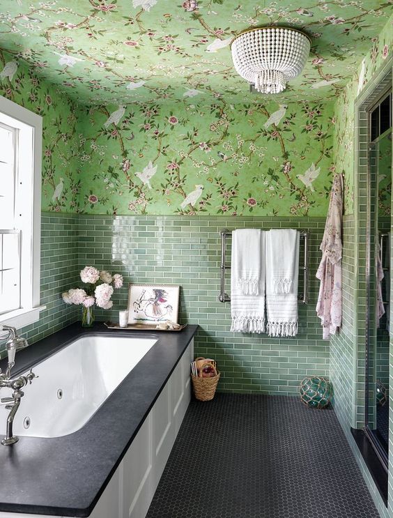 an eye catchy green bathroom with floral wallpaper and green tiles, a bathtub with paneling, a bead chandelier and some blooms