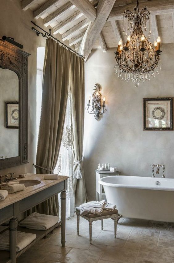 a French chic bathroom with limewashed walls, a tiled floor, a vintage clawfoot bathtub, a crystal chandelier and a vintage vanity and stools