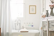 a French cottage bathroom clad with white tiles, a planked floor, a modern bathtub, a glass storage unit and artwork