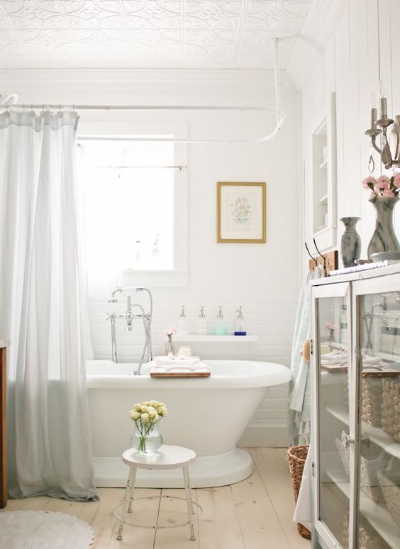 a French cottage bathroom clad with white tiles, a planked floor, a modern bathtub, a glass storage unit and artwork