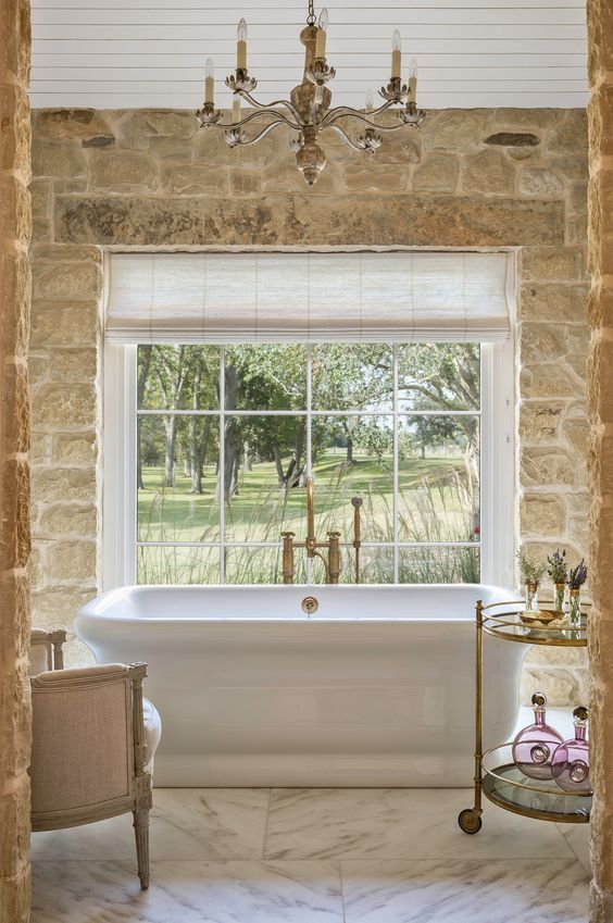 a French country style bathroom clad with marble tiles and stone, with a tub opposite the window, a vintage chandelier and vintage furniture