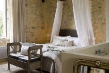 a Provencal bedroom with stone walls, a dark bed with a canopy, a wooden bench with upholstery, various textiles is cool