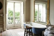 a Provence bathroom with a rough wooden plank floor, an oversized mirror in a gilded frame, a navy clawfoot bathtub, a crystal chandelier and a stained stool