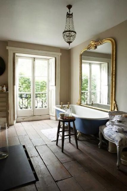 a Provence bathroom with a rough wooden plank floor, an oversized mirror in a gilded frame, a navy clawfoot bathtub, a crystal chandelier and a stained stool