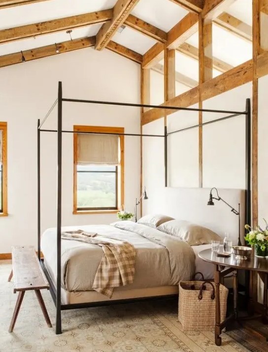 a Provence style bedroom with wooden beams, a series of windows for natural light, a metal canopy bed with grey bedding, round nightstands and a wooden bench