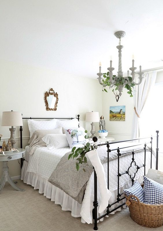 a beautiful Provence bedroom with a metal bed with neutral bedding, white and grey nightstands, greenery, a basket with pillows and a mirror in a chic frame
