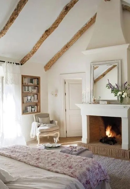 a beautiful neutral and pastel Provence bedroom with a fireplace, built-in shelves, a bed with pastel bedding, a refined chair and a lilac pouf
