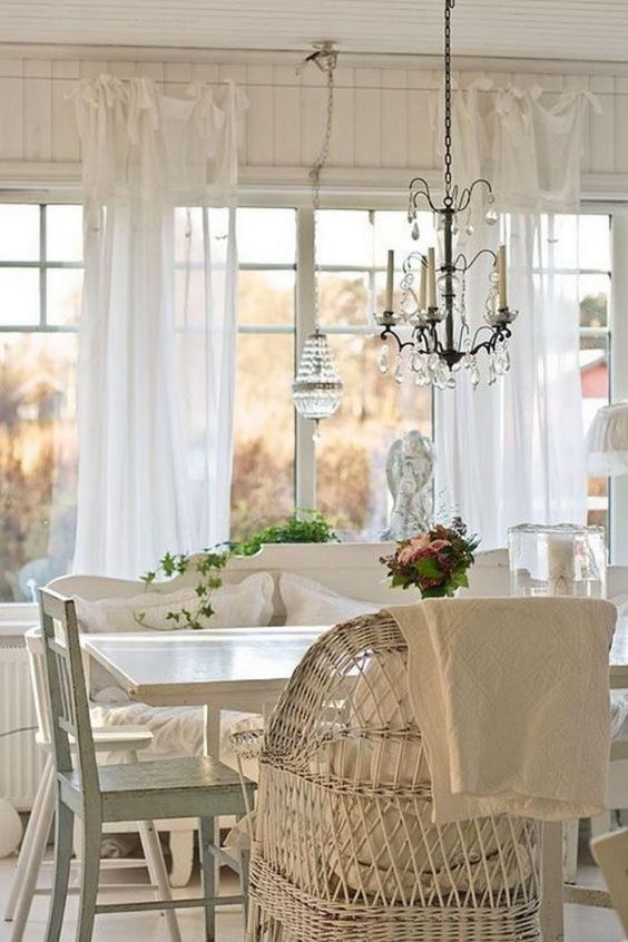 a beautiful vintage cottage sunroom in white, with wooden and wicker furniture, a crystal chandelier and greenery