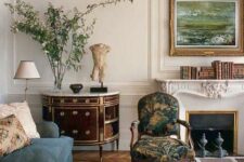 a beautiful vintage living room with stucco and a chic fireplace, a blue sofa, an elegant dresser, a printed chair and some greenery