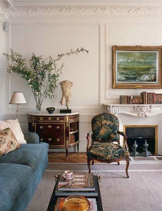 a beautiful vintage living room with stucco and a chic fireplace, a blue sofa, an elegant dresser, a printed chair and some greenery