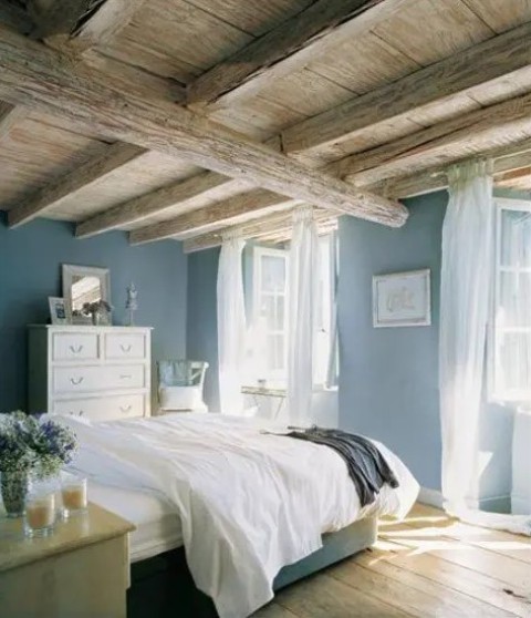 a blue Provence bedroom with wooden beams on the ceiling, a bed with neutral bedding, a white dresser, mirrors and frames and candles