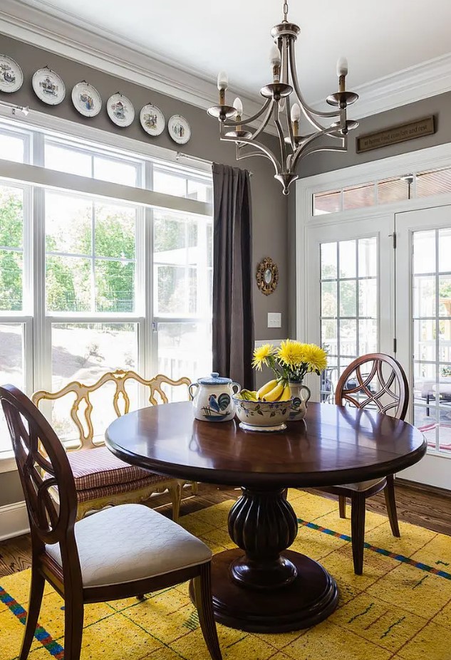 a bold vintage dining room with grey walls, a large window, a heavy round table and chairs, decorative plates and a bold yellow rug