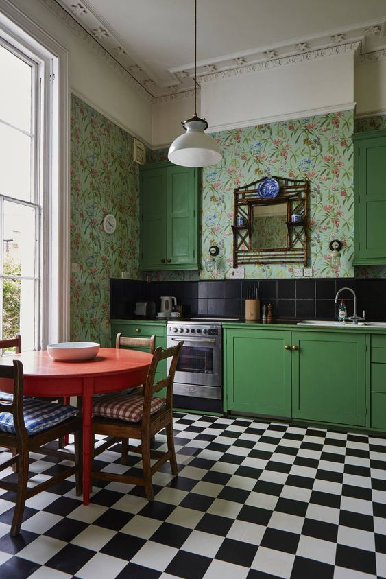 a bold vintage kitchen with bright green cabinets, bright floral wallpaper, black tiles on the backsplash, a checked floor and a round red table