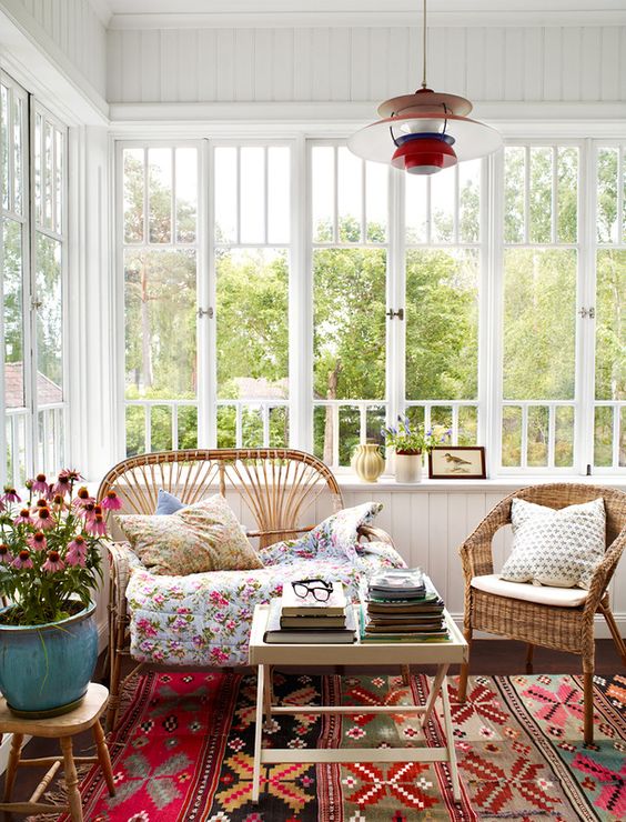 a bright vintage sunroom with a colorful rug, wicker furniture and floral pillows, a potted plant and a mini table