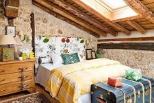 a catchy and colorful vintage attic bedroom with a floral bed, bold bedidng and rugs, a vintage blue chest and stone walls
