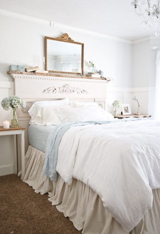 a chic Provencal bedroom with white paneling, a bed with a refined headboard, blue and white bedding, books and a mirror, a crystal chandelier