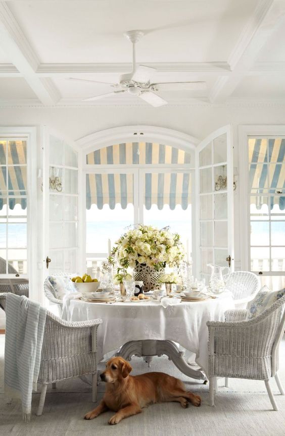 a classic vintage sunroom done in all neutrals, with a white wicker furniture set, potted blooms and neutral textiles