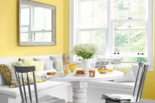 a colorful dining nook with yellow walls, a white corner seat, a white table, grey chairs and grey and yellow pillows