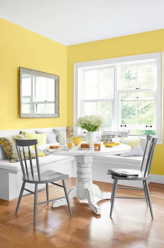 a colorful dining nook with yellow walls, a white corner seat, a white table, grey chairs and grey and yellow pillows