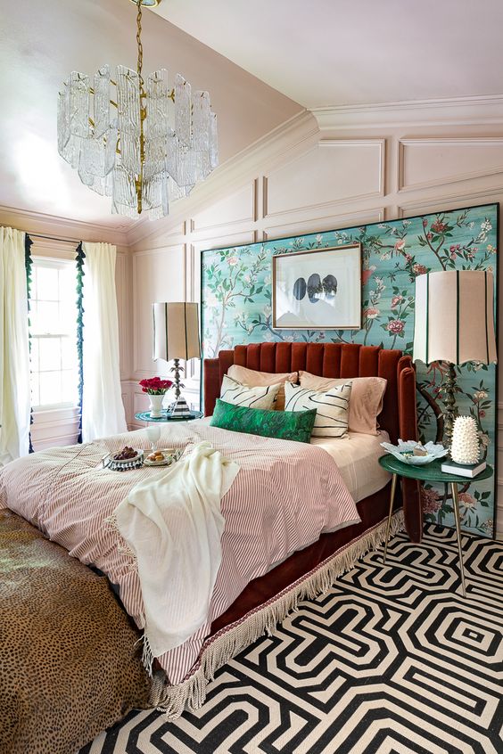 a colorful vintage bedroom with paneling, a bright floral wall, a burgundy bed, colorful bedding and a crystal chandelier