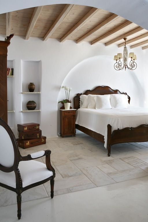 a contrasting Provence bedroom with a dark-stained in a niche, built-in shelves, dark suitcases and a dark bookcase, a chic chandelier and a chair