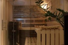 a cozy tine home sauna with wooden benches, a stone accent wall and some intimate light plus a glass wall