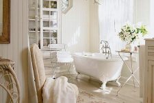 a creamy Provencal bathroom clad with wooden planks, with a glass storage unit, a clawfoot bathtub, printed rugs and shabby chic chairs and stools