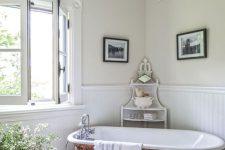 a lovely French country chic bathroom with wallpaneling, wooden beams, a vintage chandelier, a clawfoot bathtub, a wooden bench and some blooms