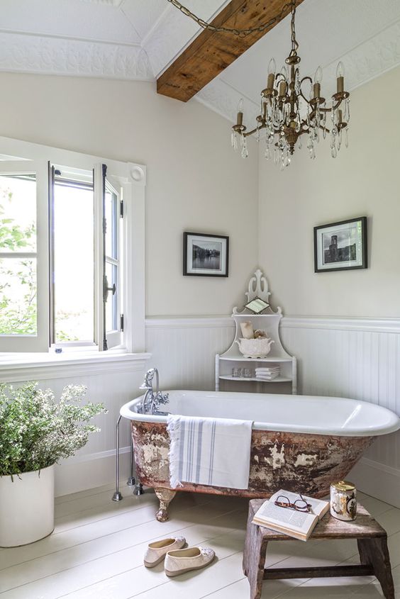 a lovely French country chic bathroom with wallpaneling, wooden beams, a vintage chandelier, a clawfoot bathtub, a wooden bench and some blooms