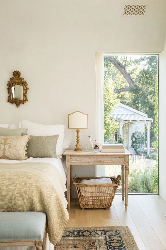 a lovely Provencal bedroom with a whitewashed table as a nightstand, a bed with earthy tone bedding, floor to ceiling windows and a printed rug