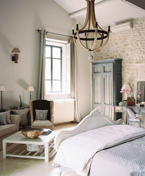 a lovely Provence bedroom with a stone accent wall, a refined white bed, grey seating furniture, a blue wardrobe and a vanity, a wooden chandelier and chic lamps