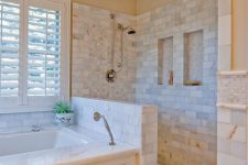 a lovely bathroom clad with marble tiles, with a half wall in the shower space, a bathtub clad with panels