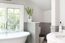 a lovely farmhouse bathroom with a tile floor, grey paneling on the walls, a vintage tub and a chic chandelier plus a half wall that separates the space