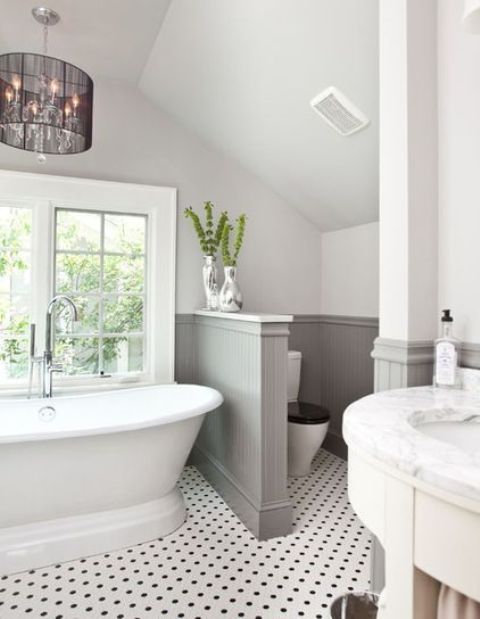 a lovely farmhouse bathroom with a tile floor, grey paneling on the walls, a vintage tub and a chic chandelier plus a half wall that separates the space