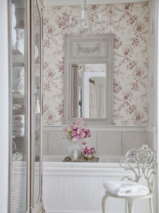 a neutral French country bathroom with floral wallpaper, white paneling, a tub clad with plants, a vintage mirror and a crystal chandelier
