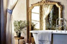 a neutral Provence bathroom with buttermilk walls, a clawfoot bathtub, a large mirror in a gilded frame, semi-sheer curtains and a basket for storage