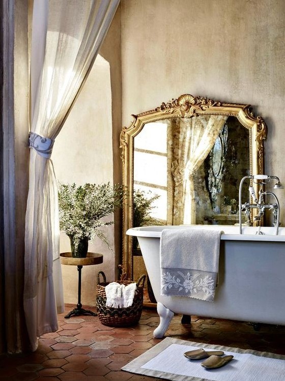 a neutral Provence bathroom with buttermilk walls, a clawfoot bathtub, a large mirror in a gilded frame, semi sheer curtains and a basket for storage