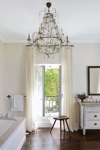 a neutral Provence bathroom with tan walls, a modern tub clad with paneling, a vintage vanity, a large crystal chandelier and a stained stool