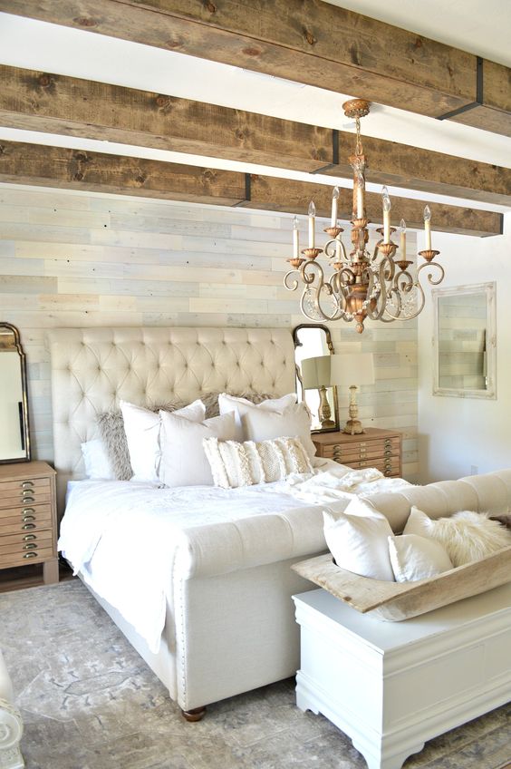 a neutral Provence bedroom with a catchy pastel accent wall, wooden beams on the ceiling, an upholstered neutral bed with neutral bedding, a white storage chest and a chic chandelier
